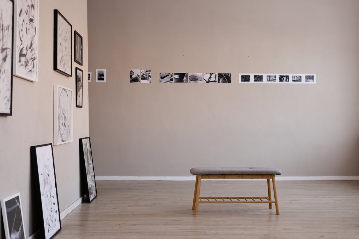 Empty art gallery displaying black and white framed photographs and prints on beige walls with wooden bench in foreground, showcasing contemporary minimalist exhibition setup